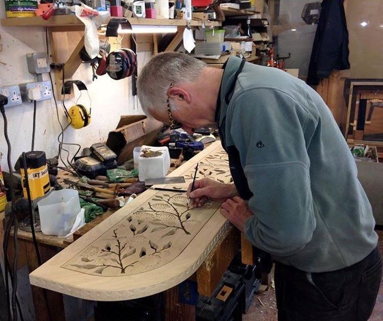 A cabinetmaker hand-working the tree motif into a curved timber frieze at the bench in the Stroud Furniture Makers workshop