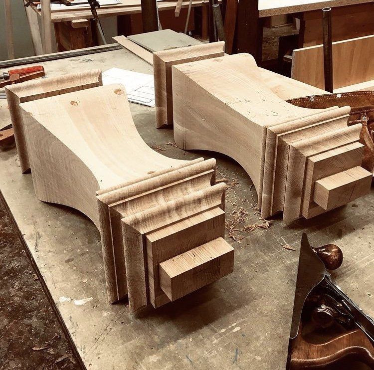 Two solid-timber pedestal bases mid-shape on the bench with a hand plane and shavings, in the Stroud Furniture Makers workshop